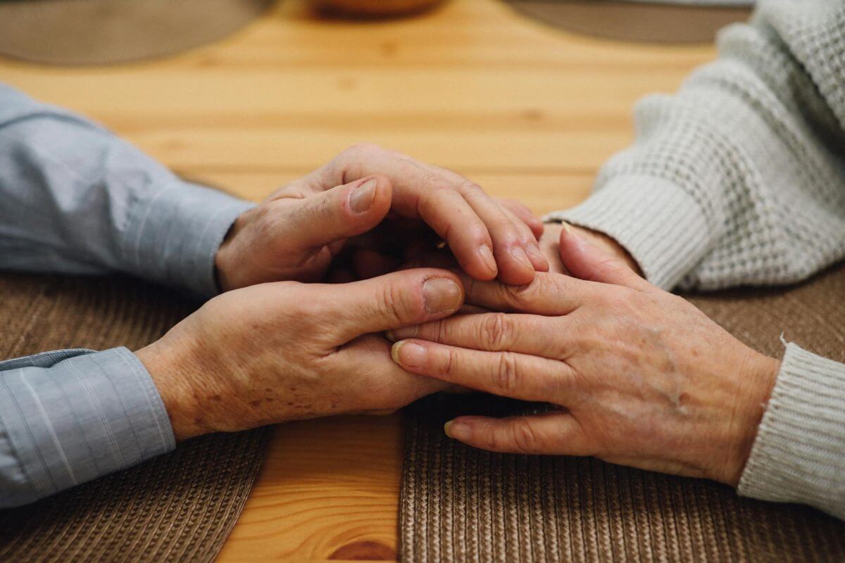 Two people sit at a table, gently holding each other's hands in a comforting gesture, seeking guidance from Financial Planner Pinsker Wealth.