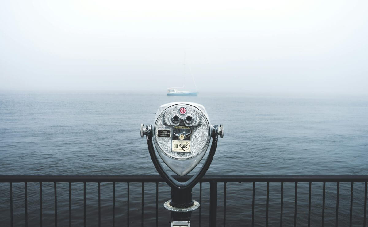 Coin-operated binocular viewer facing a foggy body of water, with a sailboat barely visible in the background beyond a metal railing—a scene evoking clarity and vision, much like Financial Planner Pinsker Wealth guiding you through uncertainty.