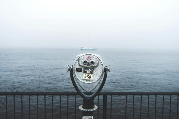 Coin-operated binocular viewer facing a foggy body of water, with a sailboat barely visible in the background beyond a metal railing—a scene evoking clarity and vision, much like Financial Planner Pinsker Wealth guiding you through uncertainty.