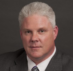 A middle-aged man with short gray hair, Financial Planner at Pinsker Wealth, wears a dark suit, white shirt, and striped tie while posing against a plain dark background.
