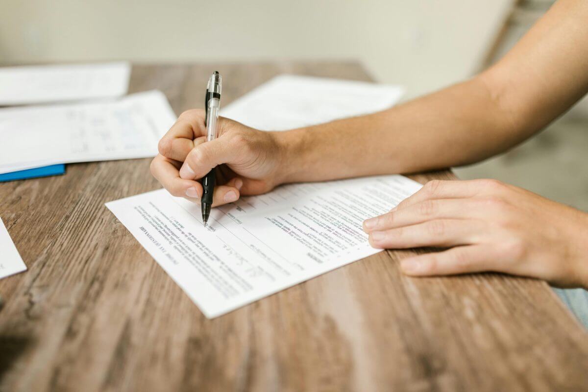 A person fills out a printed form with a pen on a wooden table, with additional papers in the background—an everyday scene for a Financial Planner at Pinsker Wealth.