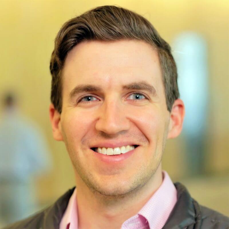 A man with light skin and brown hair smiles at the camera, wearing a light pink collared shirt and a dark jacket—capturing the approachable professionalism of Financial Planner Pinsker Wealth. The background is softly blurred.